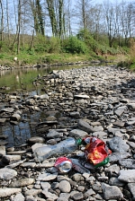 Auf dem Bild sieht man einen kleinen Fluss oder Bach mit vielen Steinen am Ufer. Das Wasser ist flach, und im Hintergrund stehen Bäume.

Vorne im Bild liegt Müll auf den Steinen: eine leere Plastikflasche, eine zerdrückte Getränkedose und eine aufgerissene Chips-Tüte.