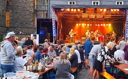 Zu sehen sind die Gäste der Parkzeit. Im Hintergrund ist die Bühne auf der eine Live Band performt. Die Gäste sitzen auf Bierbänken an Biertischen und genießen mitgebrachte Speisen und Getränke.