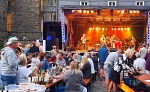 Zu sehen sind die Gäste der Parkzeit. Im Hintergrund ist die Bühne auf der eine Live Band performt. Die Gäste sitzen auf Bierbänken an Biertischen und genießen mitgebrachte Speisen und Getränke.
